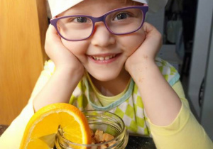 Uśmiechnięta dziewczynka w czapce kucharskiej siedzi przed gotowym deserem w słoiczku, ozdobionym plastrem pomarańczy.